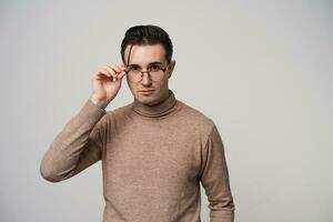 retrato do bonita jovem Sombrio cabelos masculino hipster dentro óculos olhando para Câmera com calma face enquanto em pé sobre branco fundo, vestindo bege pescoço enrolado suéter foto