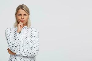 retrato do sério concentrado Loiras jovem mulher desgasta polca ponto camisa mantém mãos guardada, parece pensativo e pensando isolado sobre branco fundo foto