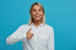 retrato do feliz lindo Loiras jovem mulher desgasta polca ponto camisa parece confiante e mostra polegares acima gesto isolado sobre azul fundo foto