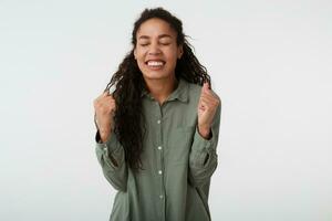 Boa olhando jovem alegre Sombrio cabelos encaracolado senhora com Sombrio pele guardando dela olhos fechadas e levantando mãos enquanto alegria sobre algo, isolado sobre branco fundo foto