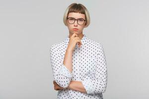 retrato do pensativo lindo Loiras jovem mulher desgasta polca ponto camisa e óculos parece sério mantém mãos guardada isolado sobre branco fundo foto