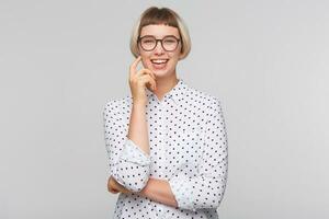 retrato do feliz lindo Loiras jovem mulher desgasta polca ponto camisa e óculos parece alegre, mantém mãos guardada e rindo isolado sobre branco fundo foto