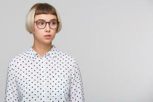 retrato do pensativo atraente Loiras jovem mulher desgasta polca ponto camisa e óculos parece sério e parece para a lado isolado sobre branco fundo foto