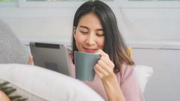 bela mulher asiática sorridente atraente usando tablet segurando uma xícara quente de café ou chá enquanto estava deitado no sofá quando relaxa na sala de estar em casa. mulheres de estilo de vida no conceito de casa.