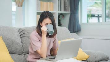 mulher asiática freelance jovem negócios trabalhando no laptop, verificando as mídias sociais e bebendo café enquanto estava deitado no sofá quando relaxava na sala de estar em casa. mulheres de estilo de vida no conceito de casa.