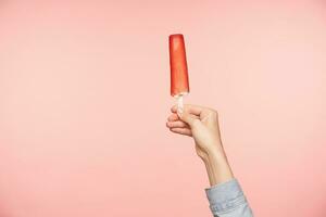 horizontal foto do jovem mulher mão segurando sorvete em bastão com vermelho Esmalte, isolado sobre Rosa fundo. Comida fotografia e humano mãos conceito