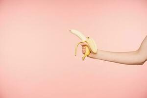 lado Visão do elevado bem cuidado feminino mão com nu manicure segurando descascado banana enquanto posando sobre Rosa fundo, indo para ter saudável lanche foto
