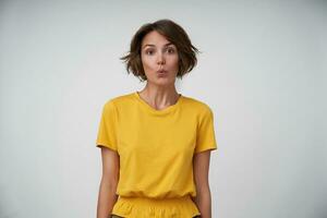 retrato do jovem morena mulher com casual Penteado vestindo amarelo camiseta posando sobre branco fundo com Hans abaixo, olhando às Câmera positivamente e perseguindo dela lábios foto