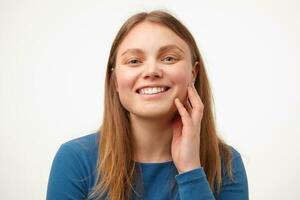 feliz jovem adorável grandes cabelos fêmea com casual Penteado tocante dela bochecha com elevado mão e sorridente alegremente às Câmera, isolado sobre branco fundo foto