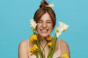 estúdio tiro do jovem alegre ruiva fêmea com pão Penteado franzindo a testa alegremente dela face enquanto sorridente largamente, isolado sobre azul fundo com grupo do flores foto