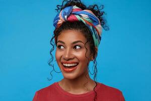 estúdio foto do jovem adorável morena encaracolado senhora com colhido cabelo vestido dentro colori na moda roupas olhando alegremente a parte, de lado com Largo sorriso, isolado sobre azul fundo