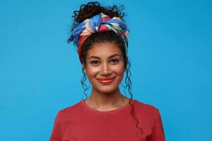 retrato do satisfeito jovem adorável Sombrio cabelos encaracolado mulher com colhido cabelo olhando positivamente às Câmera com encantador sorriso, isolado sobre azul fundo foto