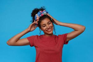 satisfeito jovem alegre morena encaracolado mulher com colhido cabelo segurando mãos em dela cabeça e sorridente amplamente com fechadas olhos, isolado sobre azul fundo foto