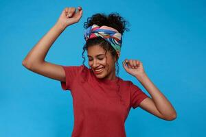 Boa olhando jovem bonita morena encaracolado mulher com colhido cabelo sorridente alegremente com fechadas olhos enquanto dançando com elevado mãos, posando sobre azul fundo foto