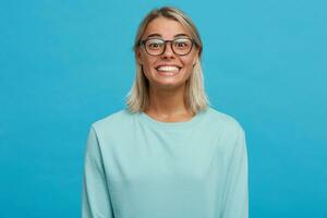 Tiros na Cabeça do engraçado feliz feliz Loiras jovem menina dentro copos, amplamente sorrisos, brilhando a partir de felicidade, completamente satisfeito, vestido dentro luz azul grandes manga camiseta, em uma azul fundo foto