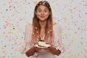 feliz animado jovem mulher com grandes tingido pastel Rosa cabelo desgasta polca ponto Rosa vestir segurando prato com peça do bolo com vela e tendo festa isolado sobre branco fundo com confete foto