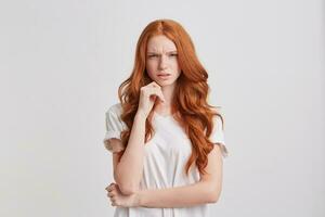 fechar-se do chateado infeliz ruiva jovem mulher com grandes ondulado cabelo e sardas desgasta à moda t camisa mantém mãos guardada e parece Bravo isolado sobre branco fundo foto