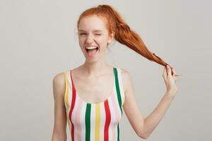 retrato do alegre atraente jovem mulher com vermelho cabelo e sardas dentro listrado topo segurando dela rabo de cavalo e piscando isolado sobre branco fundo foto