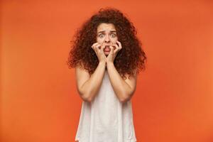 receoso olhando mulher, menina com encaracolado gengibre cabelo. vestindo branco ombros caídos blusa. tocante dela face. tem medo em dela face. assistindo às a Câmera horrorizado, isolado sobre laranja fundo foto