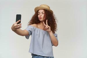 Adolescência garota, feliz mulher com encaracolado gengibre cabelo. vestindo listrado ombros caídos blusa e chapéu. levando uma selfie em uma Smartphone, mostrando Paz placa e beijo. ficar de pé isolado sobre branco fundo foto