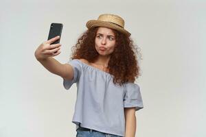 Adolescência garota, feliz olhando mulher com encaracolado gengibre cabelo. vestindo listrado ombros caídos blusa e chapéu. levando uma selfie em uma Smartphone e fazendo beicinho lábios. ficar de pé isolado sobre branco fundo foto