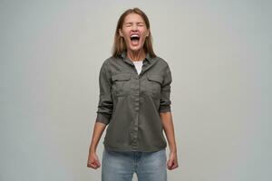 retrato do atraente, adulto menina com Castanho grandes cabelo, piercing. vestindo cinzento camisa e jeans. grito louvar com fechadas olhos, aperto dela punhos dentro raiva. ficar de pé isolado sobre cinzento fundo foto