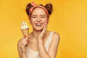 retrato do atraente, adorável menina com dois pãezinhos. vestindo branco tanque topo e vermelho dotado elástico de cabelo. segurando gelo creme, pegue dela nariz sujo dentro gelo creme, sorriso. ficar de pé isolado sobre amarelo fundo foto