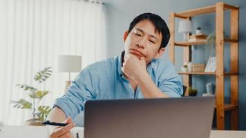 freelance asiático cara casual wear usando laptop on-line na sala de estar no escritório em casa. trabalho em casa, trabalho a distância, educação a distância, distanciamento social, quarentena para prevenção do vírus corona.