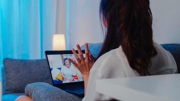 Ásia estudante intercâmbio feminino usando videochamada de laptop falando com a família enquanto trabalhava em casa na sala de estar à noite. auto-isolamento, distanciamento social, quarentena para coronavírus no próximo normal. foto