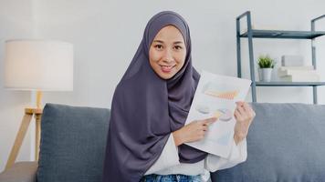 ásia senhora muçulmana usar hijab usar computador laptop conversar com colegas sobre o relatório de venda em uma reunião de videochamada enquanto trabalha remotamente em casa na sala de estar. distanciamento social, quarentena para o vírus corona.