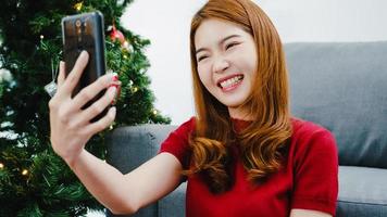 jovem mulher asiática usando videochamada de telefone inteligente falando com casal com caixa de presente de Natal, árvore de Natal decorada com enfeites na sala de estar em casa. festival de férias de Natal e ano novo.
