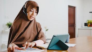 A senhora muçulmana asiática usa fone de ouvido usando tablet digital e fala com colegas sobre o relatório de venda em videoconferência enquanto trabalha em casa na cozinha. distanciamento social, quarentena para o vírus corona.