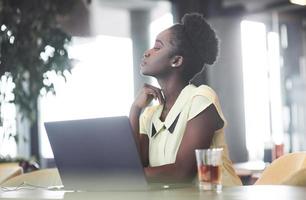 uma jovem afro-americana com cabelo escuro encaracolado pensando em um laptop em um café