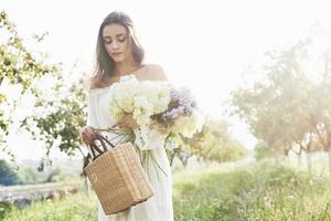 uma linda jovem em um vestido branco leve e um buquê de flores de verão faz um belo dia no jardim