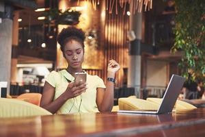 linda jovem mulher de negócios afro-americanos esperando por sua amiga em um café, telefone e laptop. foto