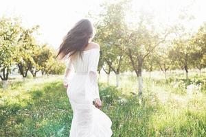 uma jovem com um vestido longo branco está caminhando no jardim. lindo pôr do sol através das folhas das árvores foto