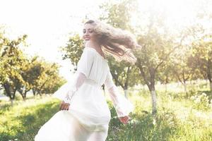 uma jovem com um vestido longo branco está caminhando no jardim. lindo pôr do sol através das folhas das árvores foto