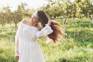 uma jovem com um vestido longo branco está caminhando no jardim. lindo pôr do sol através das folhas das árvores foto