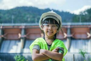 retrato do uma pequeno menina engenheiro vestindo uma verde colete e branco capacete sorridente alegremente em a fundo do a barragem. conceitos do de Meio Ambiente Engenharia, renovável energia e amor do natureza. foto