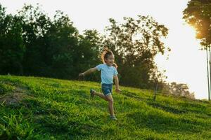 feliz pequeno menina corrida em a Relva dentro a verão parque. pequeno menina corrida através a Prado com pôr do sol claro. foto