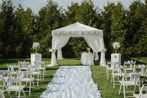 Casamento cerimônia dentro a jardim. uma branco letreiro decorado com flores, uma branco cetim tapete e muitos branco cadeiras decorado com fitas e flores uma luxuoso Casamento cerimônia. foto