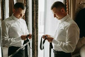 jovem homem segurando uma gravata dentro dele mãos dentro dele sala, perto a hotel quarto espelho, fechar-se foto. a noivo é preparando para a Casamento cerimônia foto
