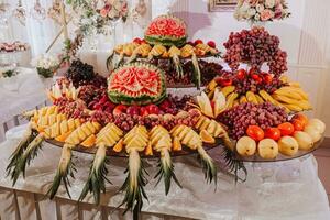 mesa com vários fruta decorações. Melancia escultura, abacaxi fatias, uvas, laranja foto