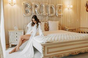 jovem encaracolado menina com grandes cabelo dentro luxuoso quarto sentado em cama dentro branco aberto vestir e preparando para Casamento cerimônia foto