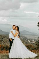 uma Casamento casal ternamente abraços dentro a montanhas. a noivo abraços a noiva de a ombros. uma linda e magnífico da noiva vestir. foto