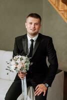 retrato do a noivo com uma ramalhete do flores dentro dele mãos, sentado em a sofá dentro dele sala. preparação do a noivo para a Casamento cerimônia. foto