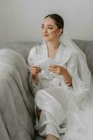 retrato do lindo menina noiva com à moda penteado e Maquiagem dentro branco roupão sentado em sofá e segurando Casamento convite. foto