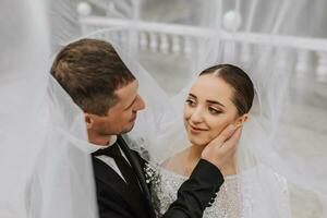 uma à moda casal do europeu recém-casados. sorridente noiva dentro uma branco vestir. a noivo, vestido dentro uma clássico Preto terno, branco camisa, debaixo uma véu. Casamento dentro natureza foto