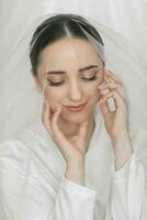 retrato do uma lindo noiva menina com à moda Penteado e Maquiagem dentro uma branco manto, debaixo uma véu, suavemente segurando dela face com dela dedos foto