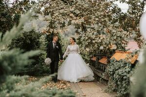 uma à moda casal do europeu recém-casados. sorridente noiva dentro uma branco vestir. a noivo é vestido dentro uma clássico Preto terno, branco camisa e gravata. Casamento dentro natureza, uma andar dentro a jardim entre alta árvores foto